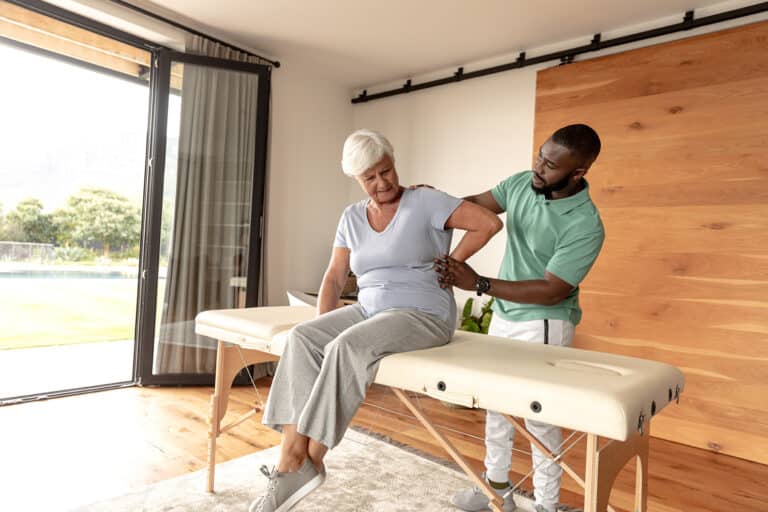 African american male physiotherapist giving back massage therapy to caucasian senior woman at home. Physiotherapy and rehabilitation concept